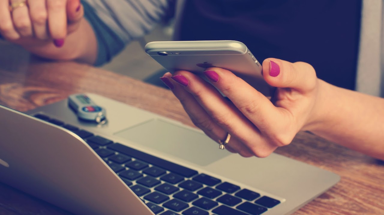 A person holding a smartphone above an open laptop.