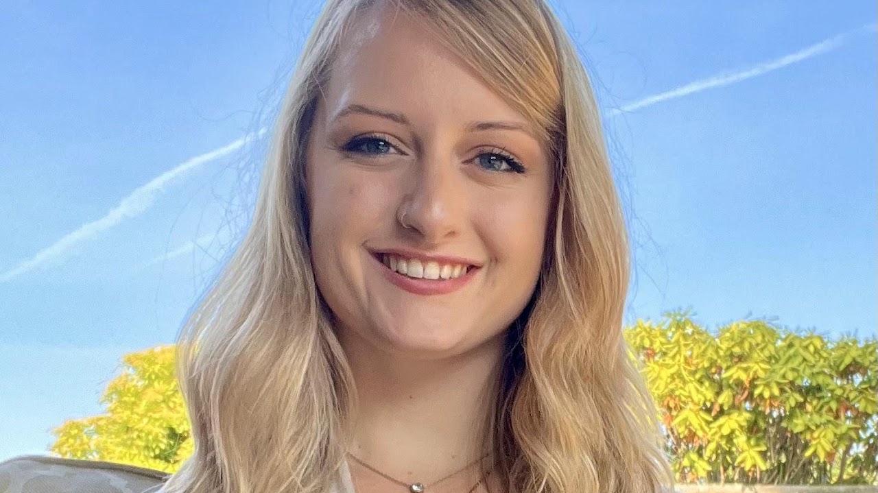 Portrait of a smiling blonde woman outdoors against a blue sky and green trees.